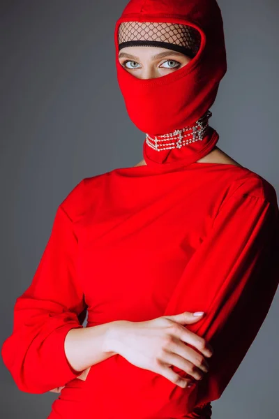 Mujer elegante en vestido rojo y pasamontañas con brazos cruzados aislados en gris - foto de stock