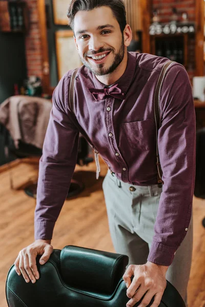 Cheerful bearded barber touching leather armchair in barbershop — Stock Photo