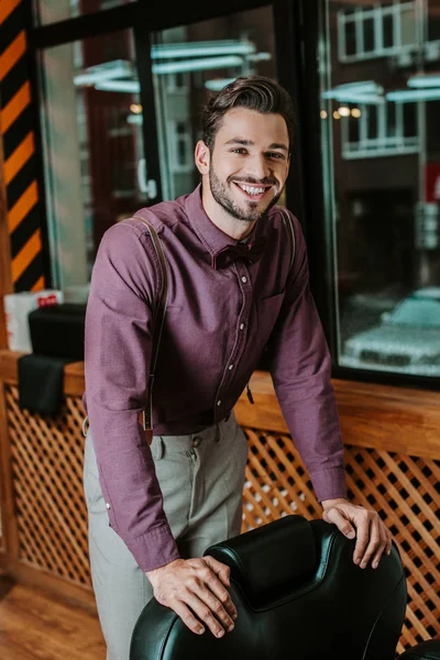 Feliz barbero barbudo tocando sillón de cuero en la barbería - foto de stock