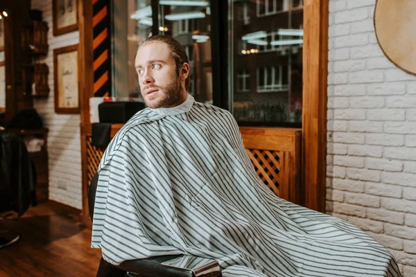Bärtiger Mann in entkleidetem Barbierumhang sitzt im Friseurladen — Stockfoto