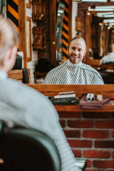 Selektiver Fokus eines glücklichen bärtigen Mannes, der im Friseurladen in den Spiegel schaut — Stockfoto