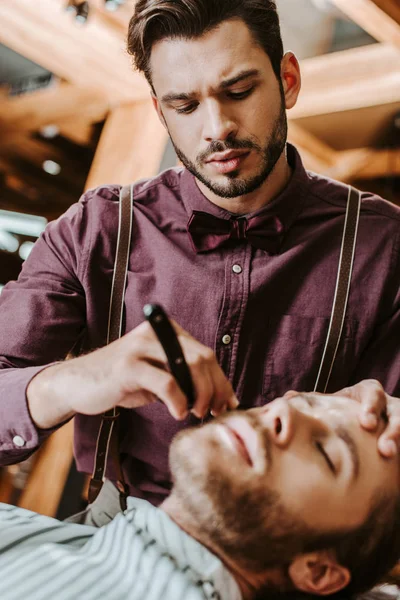 Selective focus of handsome and stylish barber shaving man — Stock Photo