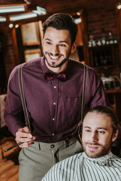 Peluquero alegre celebración de la navaja de afeitar cerca del hombre barbudo en la barbería - foto de stock