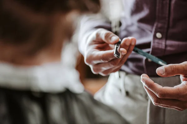 Vista cortada de barbeiro segurando navalha metálica perto do homem na barbearia — Fotografia de Stock