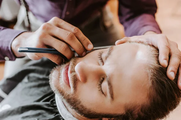 Selective focus of barber holding razor while shaving handsome man — Stock Photo