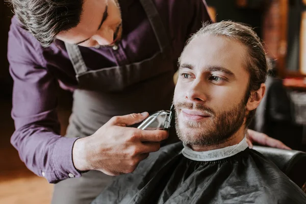 Парикмахер держит триммер во время бритья бородатый человек — стоковое фото
