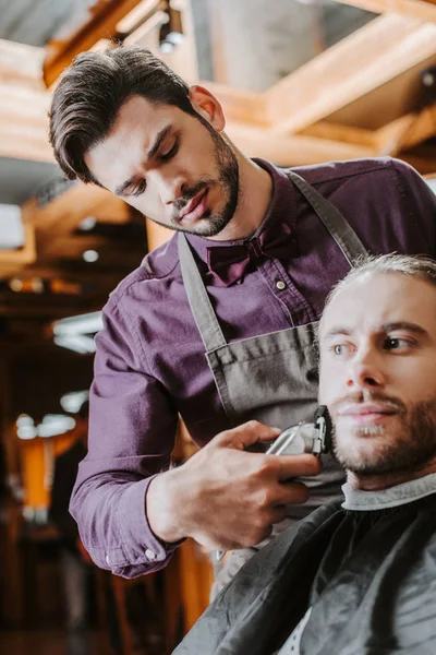 Schöner Friseur hält Trimmer, während er bärtigen Mann rasiert — Stockfoto