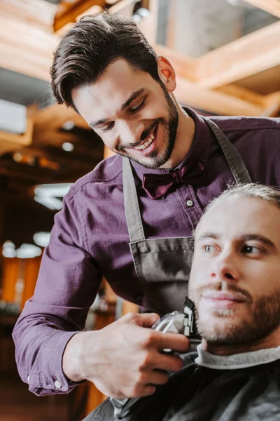 Enfoque selectivo de peluquero alegre celebración trimmer mientras afeitarse hombre barbudo - foto de stock