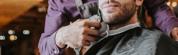 Colpo panoramico di barbiere tenuta trimmer durante la rasatura uomo barbuto — Foto stock