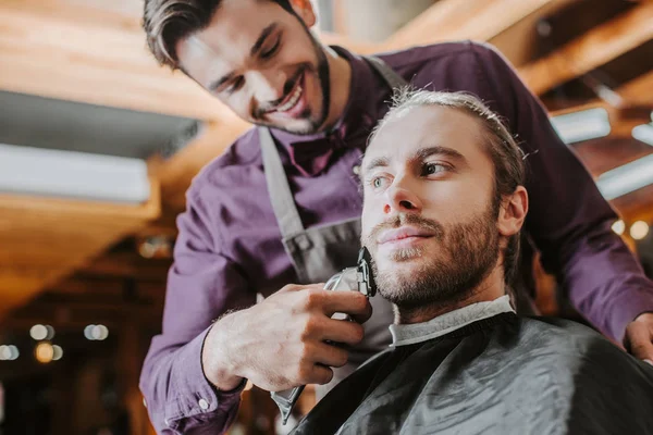 Foyer sélectif de barbier heureux tenant tondeuse tout en rasant homme barbu — Photo de stock