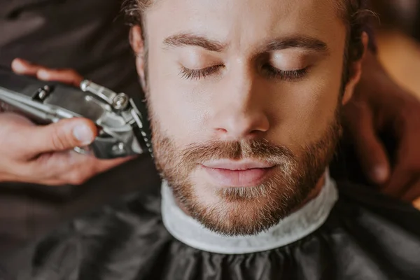 Selective focus of barber holding trimmer while shaving bearded man with closed eyes — Stock Photo