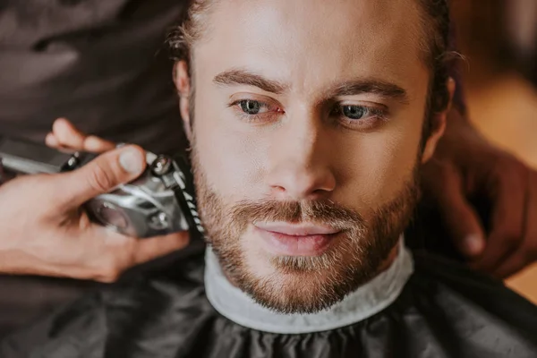 Selective focus of barber holding trimmer while shaving handsome man — Stock Photo