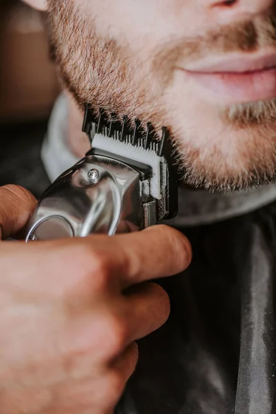 Recortado vista de peluquero celebración trimmer mientras afeitarse hombre - foto de stock
