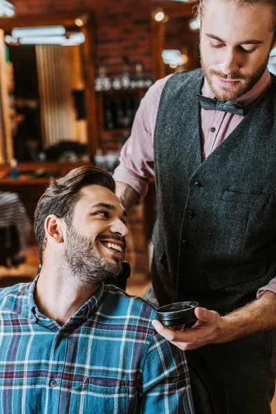 Handsome barber holding jar with black hair pomade near happy man — Stock Photo