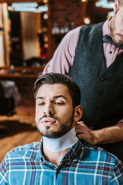 Парикмахер, крепящий воротник вокруг шеи красивого бородатого мужчины — стоковое фото