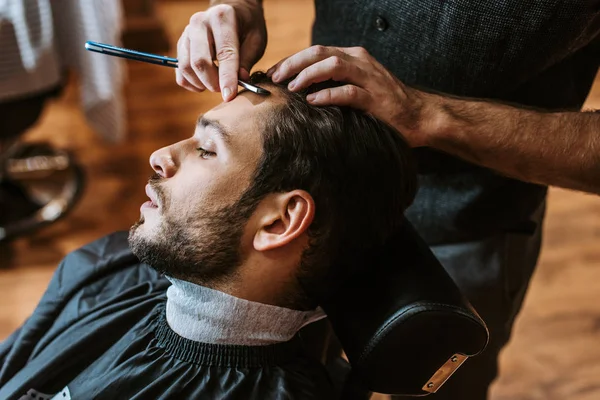 Barbeiro segurando navalha afiada ao fazer corte de cabelo para homem bonito — Fotografia de Stock