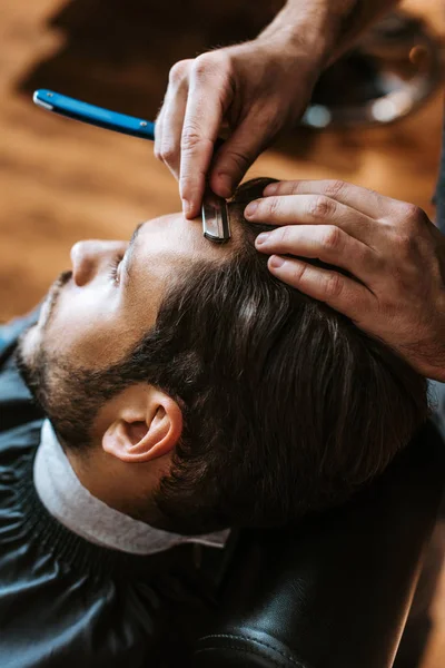 Messa a fuoco selettiva del barbiere tenendo rasoio affilato mentre si fa taglio di capelli all'uomo — Foto stock