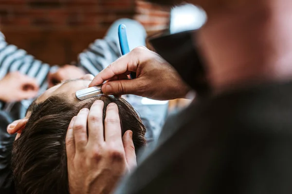 Messa a fuoco selettiva del barbiere tenuta rasoio mentre facendo taglio di capelli all'uomo — Foto stock