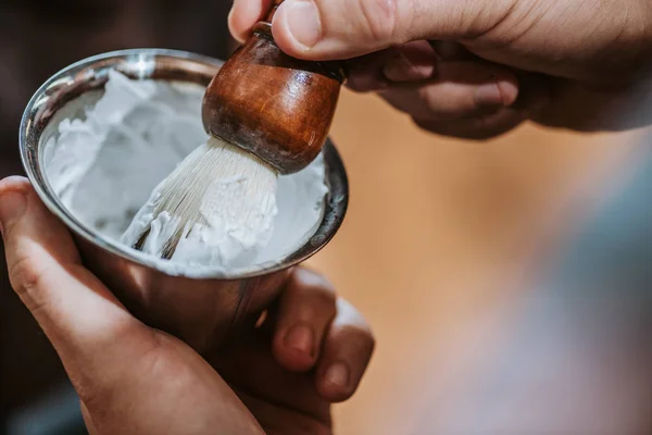 Vista ritagliata del barbiere tenendo pennello da barba vicino ciotola con crema da barba bianca — Foto stock