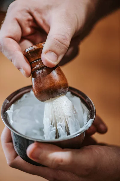 Close up de barbeiro segurando escova de barbear perto de tigela com creme de barbear branco — Fotografia de Stock