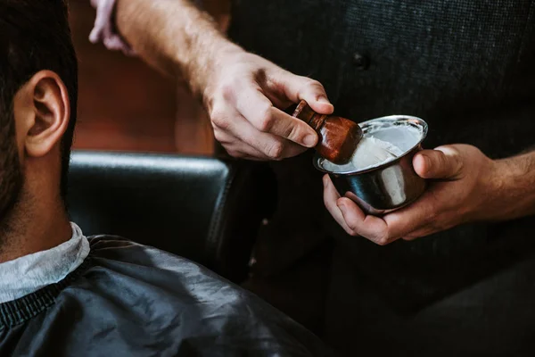 Vista recortada de peluquero sosteniendo cepillo de afeitar cerca de hombre - foto de stock