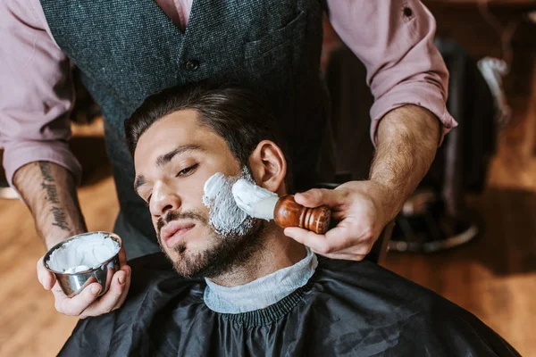Friseur trägt Rasierschaum auf Gesicht eines schönen bärtigen Mannes auf — Stockfoto