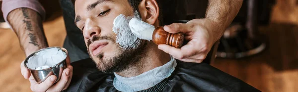 Plano panorámico de peluquero aplicando crema de afeitar en la cara del hombre barbudo guapo - foto de stock