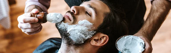 Panoramic shot of barber applying shaving cream on face of bearded man — Stock Photo
