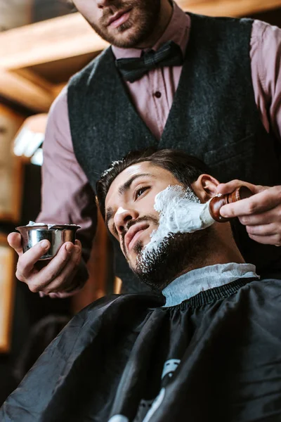 Vue à angle bas du barbier barbu appliquant de la crème à raser sur le visage de l'homme — Photo de stock