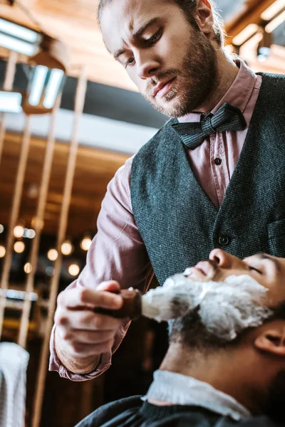Enfoque selectivo de barbero barbudo aplicación de crema de afeitar en la cara del hombre - foto de stock