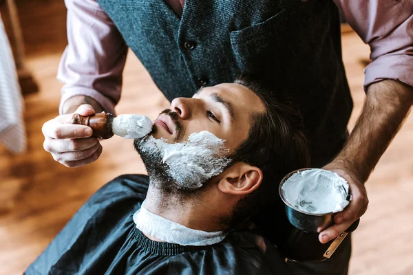 Messa a fuoco selettiva del barbiere applicare la crema da barba sul viso dell'uomo mentre tiene ciotola — Foto stock