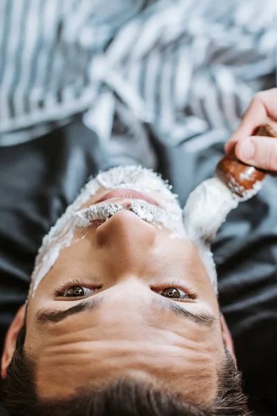 Draufsicht des Friseurs, der Rasierschaum auf das Gesicht des bärtigen Mannes aufträgt — Stockfoto