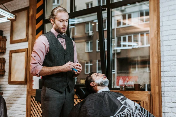Красивый парикмахер держа бритву рядом бородатый человек с кремом для бритья на лице — стоковое фото