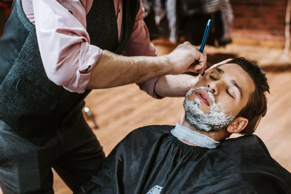 Friseur rasiert hübschen Mann mit Rasierschaum im Gesicht — Stockfoto