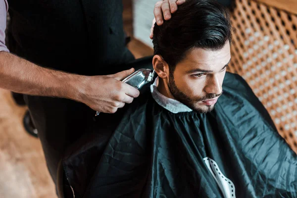 Peluquero celebración trimmer mientras peinar el cabello de hombre guapo en la barbería - foto de stock