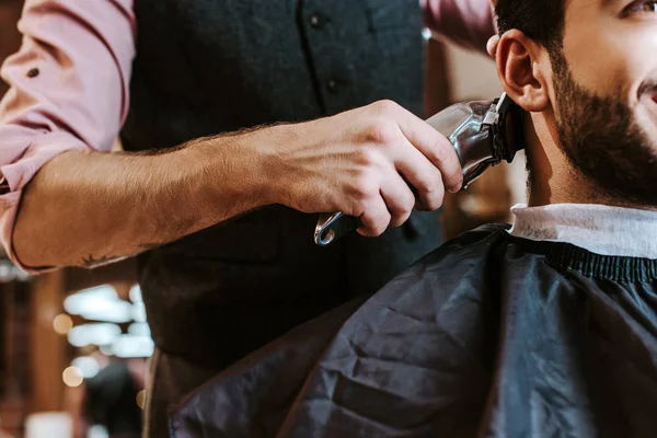 Vue recadrée du barbier tenant tondeuse tout en coiffant les cheveux de l'homme barbu dans le salon de coiffure — Photo de stock