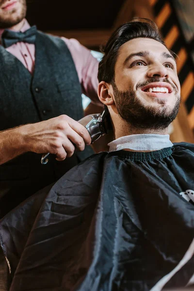 Vista angolo basso del trimmer tenuta barbiere mentre lo styling dei capelli di uomo allegro in barbiere — Foto stock