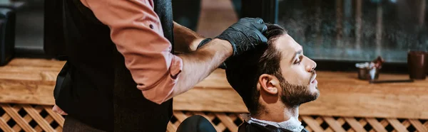 Panoramic shot of barber in black latex gloves styling hair of man — Stock Photo