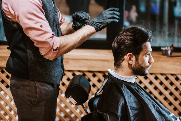 Парикмахер в черных латексных перчатках рядом с мужчиной в парикмахерской — стоковое фото