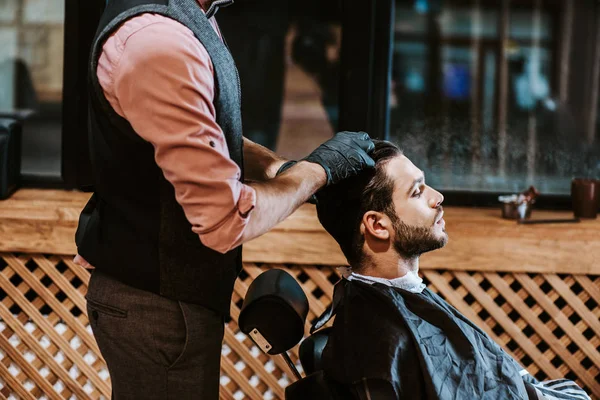 Вид сбоку бородатый мужчина сидит рядом с парикмахером в черных латексных перчатках — стоковое фото