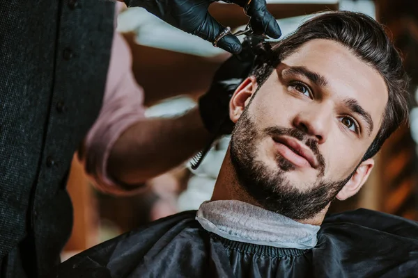 Selective focus of barber in latex gloves holding scissors while styling hair of man — Stock Photo