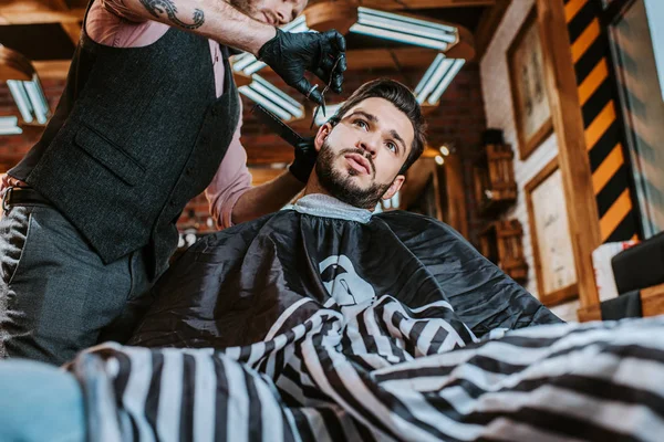 Vue à angle bas du coiffeur tatoué dans des gants en latex tenant des ciseaux tout en coiffant les cheveux de l'homme — Photo de stock