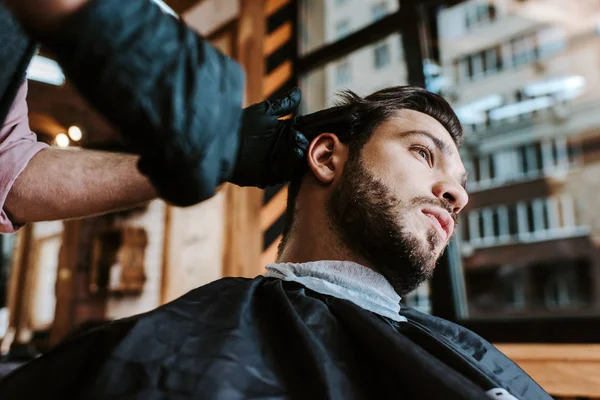 Foco seletivo do barbeiro em luvas de látex segurando pente de cabelo enquanto estiliza o cabelo do homem — Fotografia de Stock