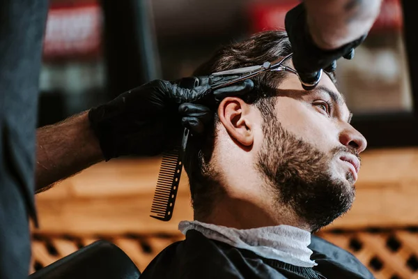 Foco seletivo do barbeiro em luvas de látex segurando pente de cabelo e tesoura enquanto estiliza o cabelo do homem — Fotografia de Stock
