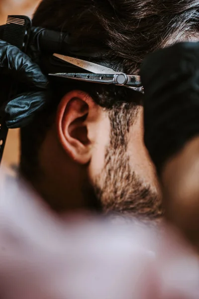 Vista recortada de peluquero en guantes de látex sosteniendo peine de pelo y tijeras mientras peina el cabello del hombre - foto de stock