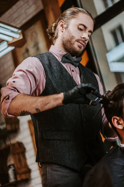 Татуйований перукар в латексних рукавичках, що тримає волосся гребінцем і ножицями під час укладання волосся людини — стокове фото