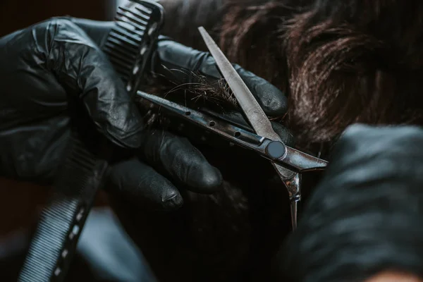 Primer plano de peluquero en guantes de látex sosteniendo peine de pelo y tijeras mientras peina el pelo del hombre - foto de stock