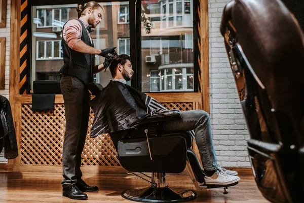 Selective focus of handsome barber styling hair of man in modern barbershop — Stock Photo