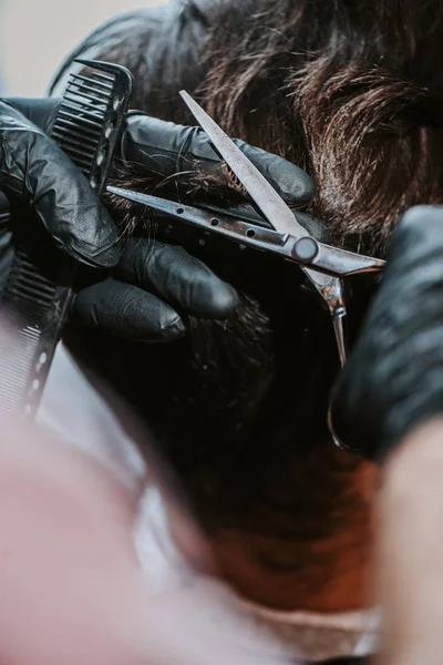 Primer plano de peluquero en guantes de látex sosteniendo peine de pelo y tijeras mientras se corta el pelo del hombre - foto de stock