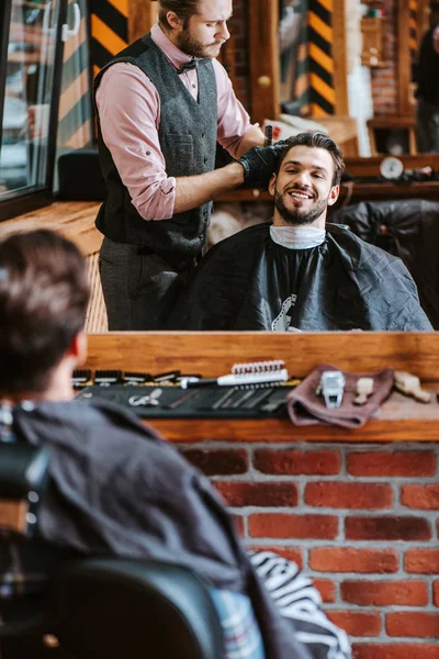 Foco seletivo de cabelo bonito barbeiro styling de homem feliz perto do espelho na barbearia — Fotografia de Stock